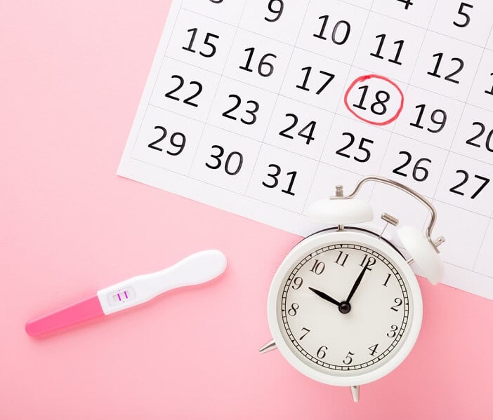 Calendar, test with two stripes and white alarm clock on pastel pink table background. Positive result. Pregnancy time beginning. Closeup. Top down view.