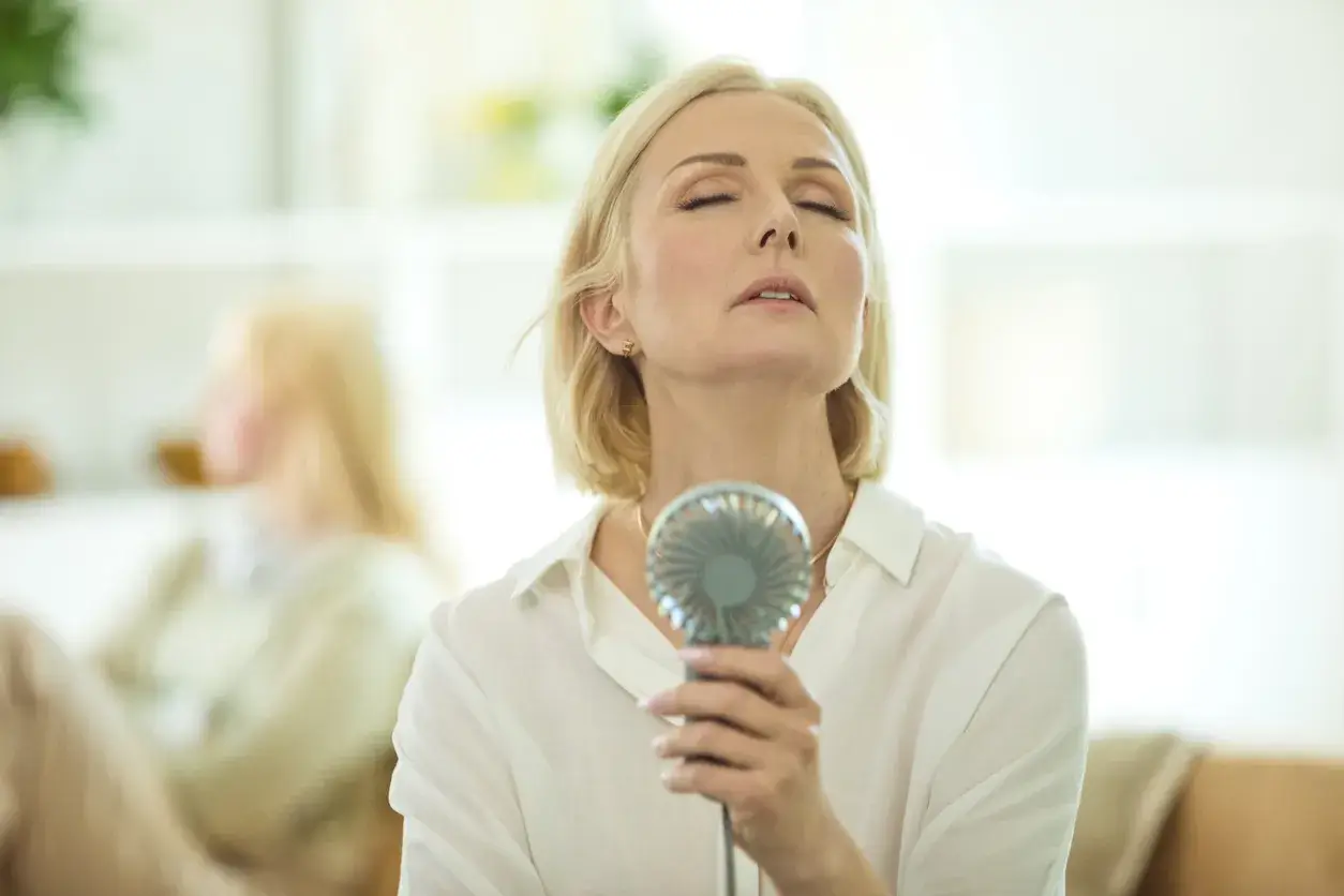 A blonde women using a fan to cool down during a hot flush due to menopause
